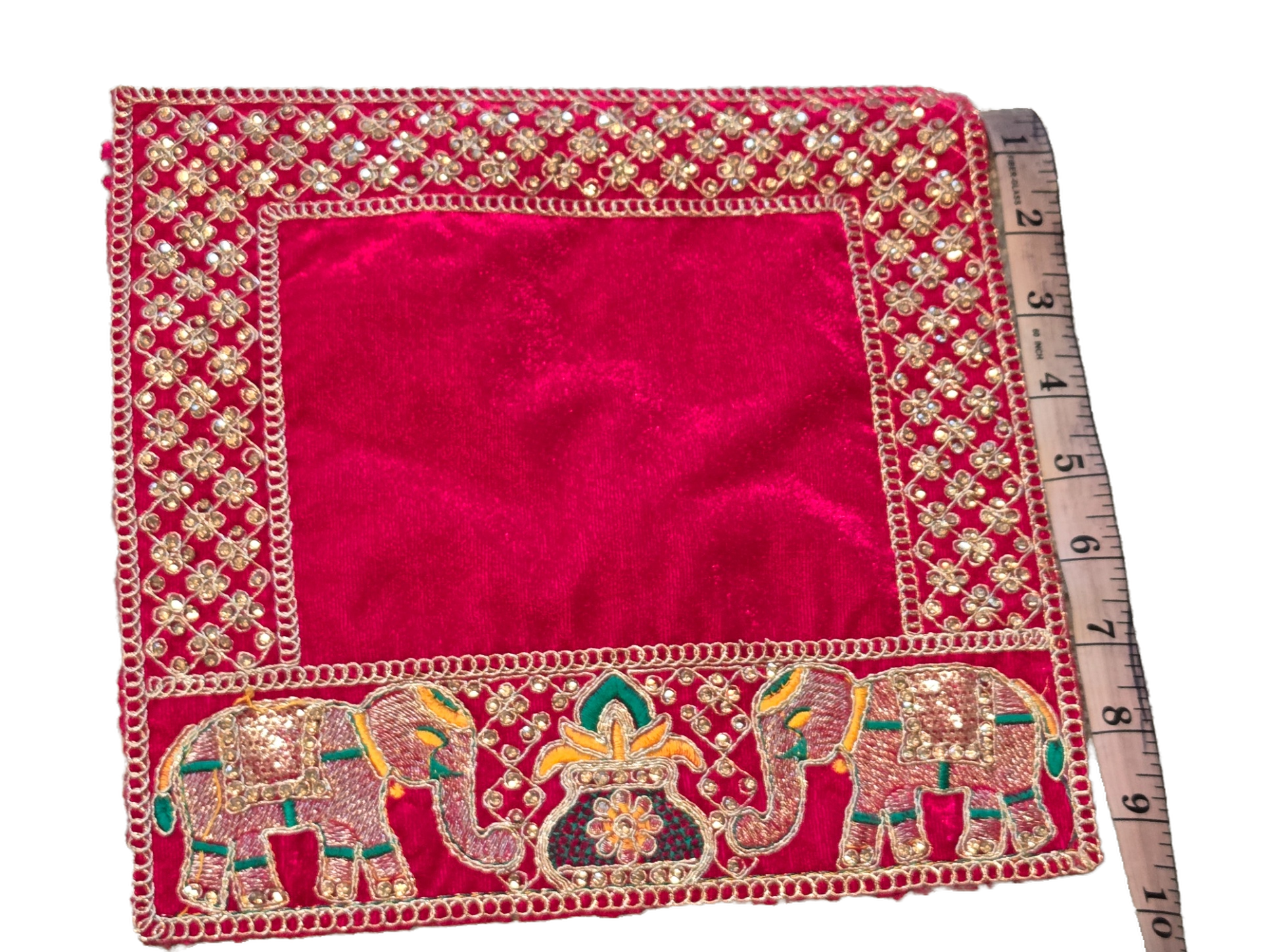 A square red velvet fabric cover with intricate gold zari embroidery, featuring elephant motifs on one side and a floral border. A wooden ruler is placed underneath to show scale.
